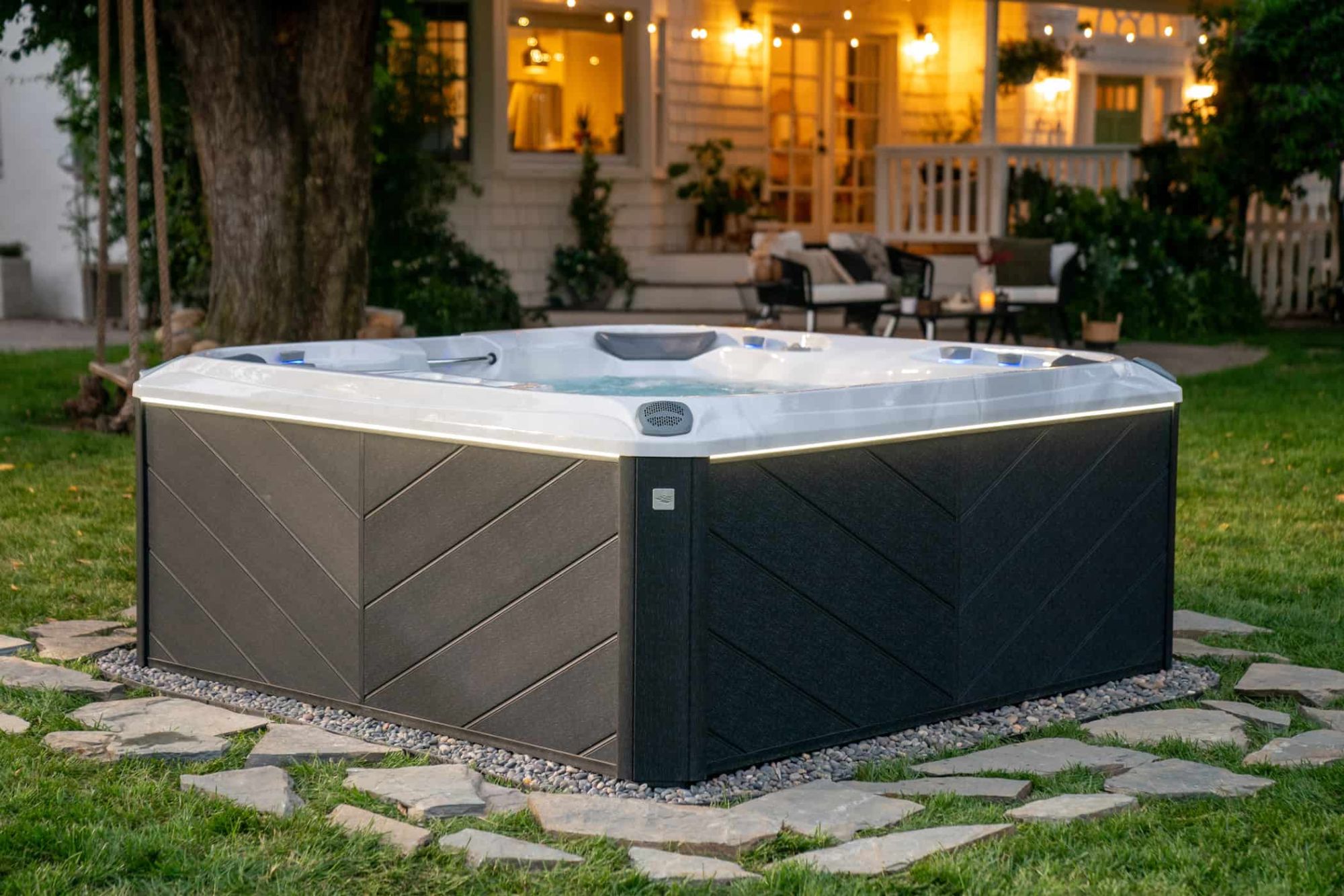 Outdoor hot tub in a yard with string lights and a lit house in the background at dusk/early evening.