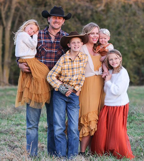 Family of six posing outdoors in fall clothing; parents with three children and a baby, all smiling at the camera.