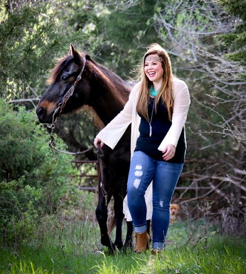 Young woman in jeans and a cardigan walking a dark brown horse through a grassy, wooded area.
