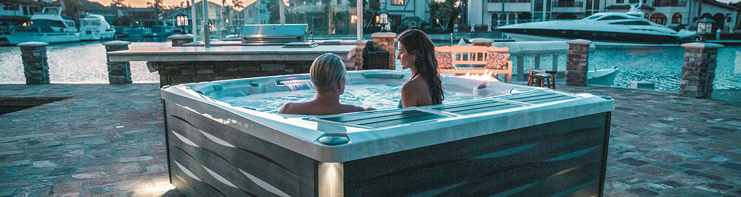 Two people sit in a rectangular hot tub on a stone patio beside a marina at dusk.