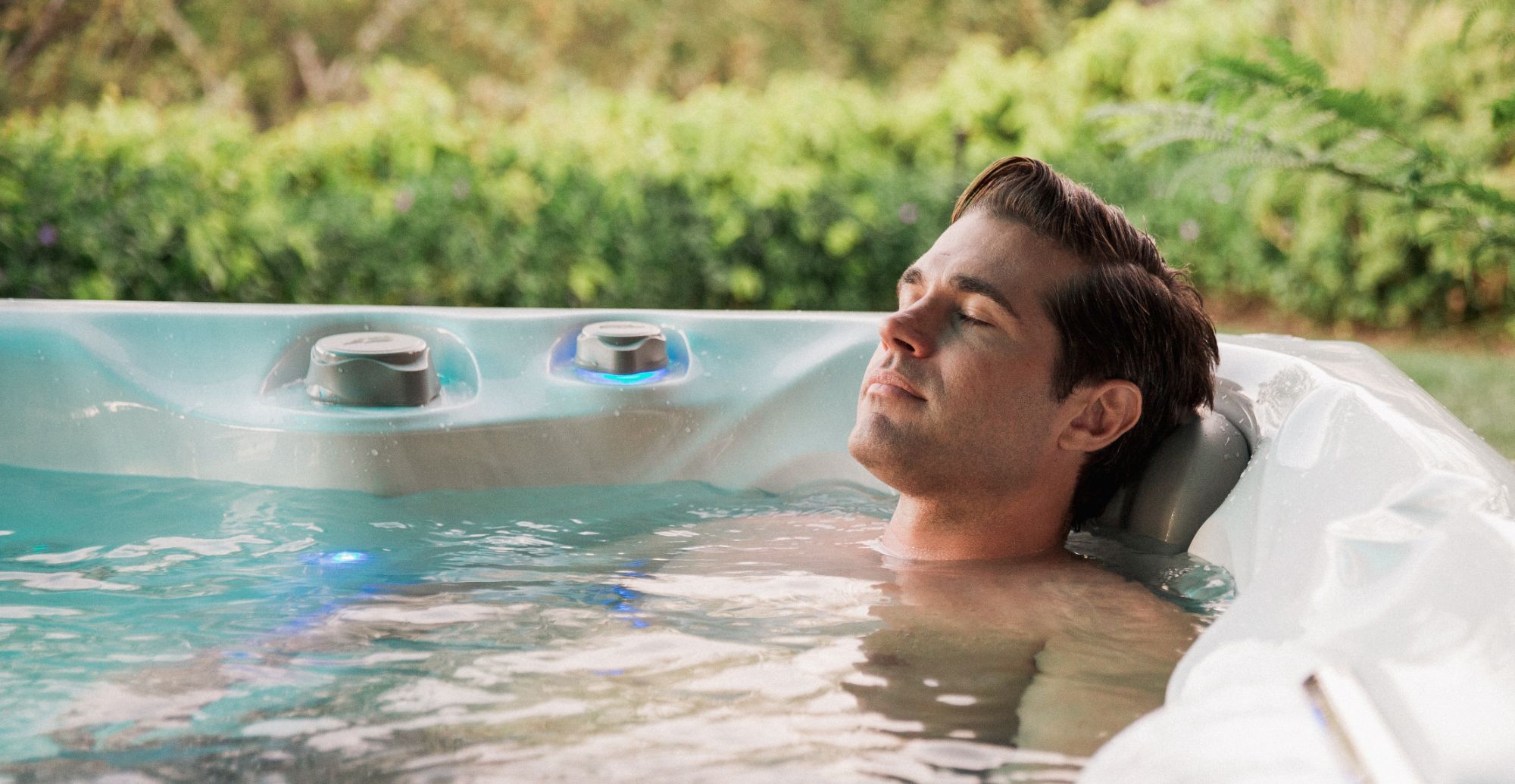 Man with eyes closed, relaxing in a bubbling outdoor hot tub, greenery in the background.