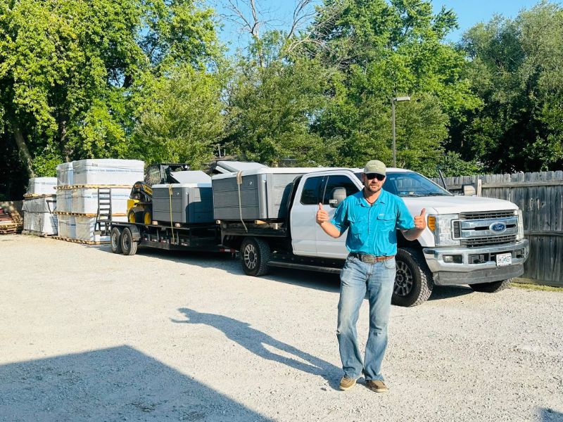 Man in a blue shirt and cap giving thumbs up beside a white pickup truck pulling a flatbed trailer loaded with large gray crates and equipment in an outdoor yard.