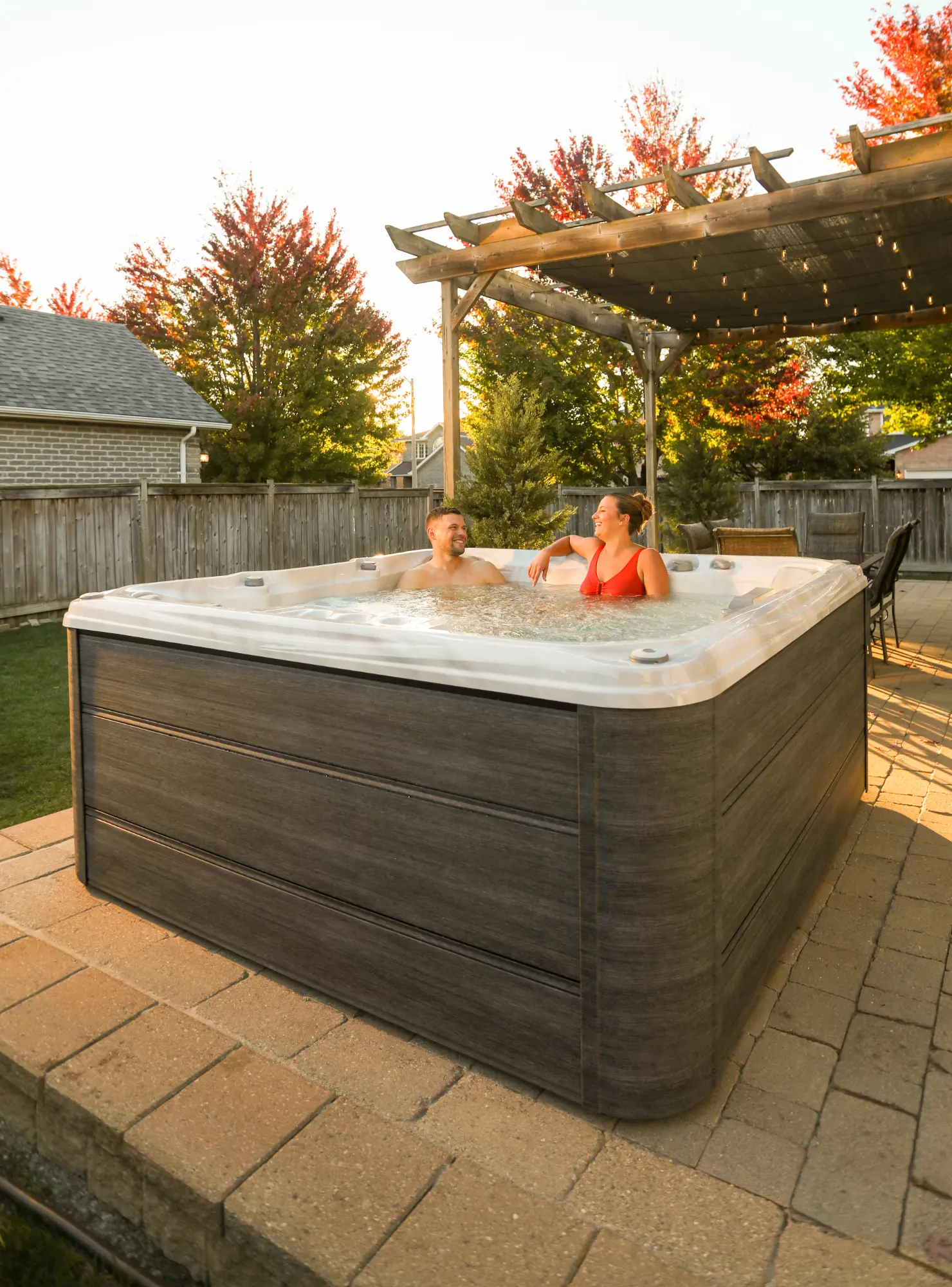 Two people relax in an outdoor hot tub in a fenced backyard at sunset, under string lights on a wooden pergola.