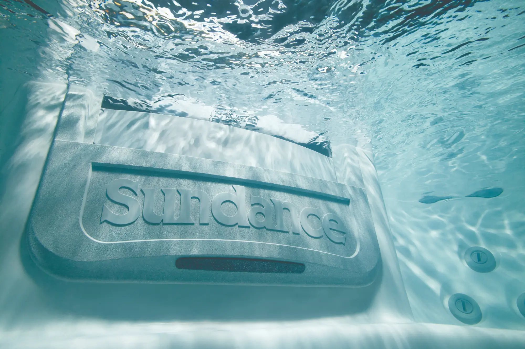 Underwater view of a metal plaque with the word 'SunDance' embossed, seen through pool water.